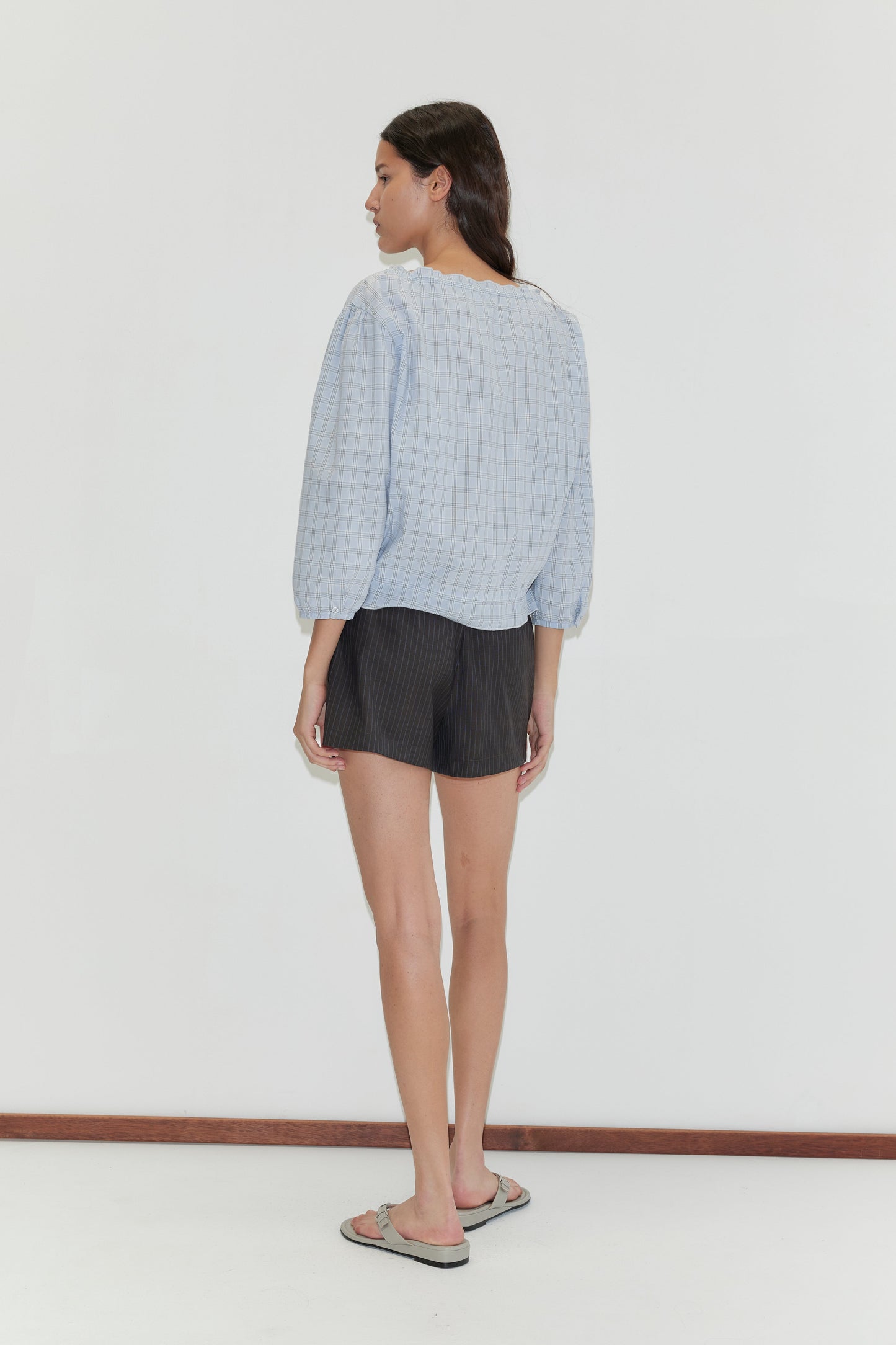 A woman faces away in a Corded Top - Powder Check by Deiji Studios, paired with black shorts and flip-flops, set against a plain white background.
