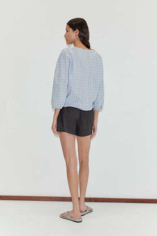 A woman faces away in a Corded Top - Powder Check by Deiji Studios, paired with black shorts and flip-flops, set against a plain white background.