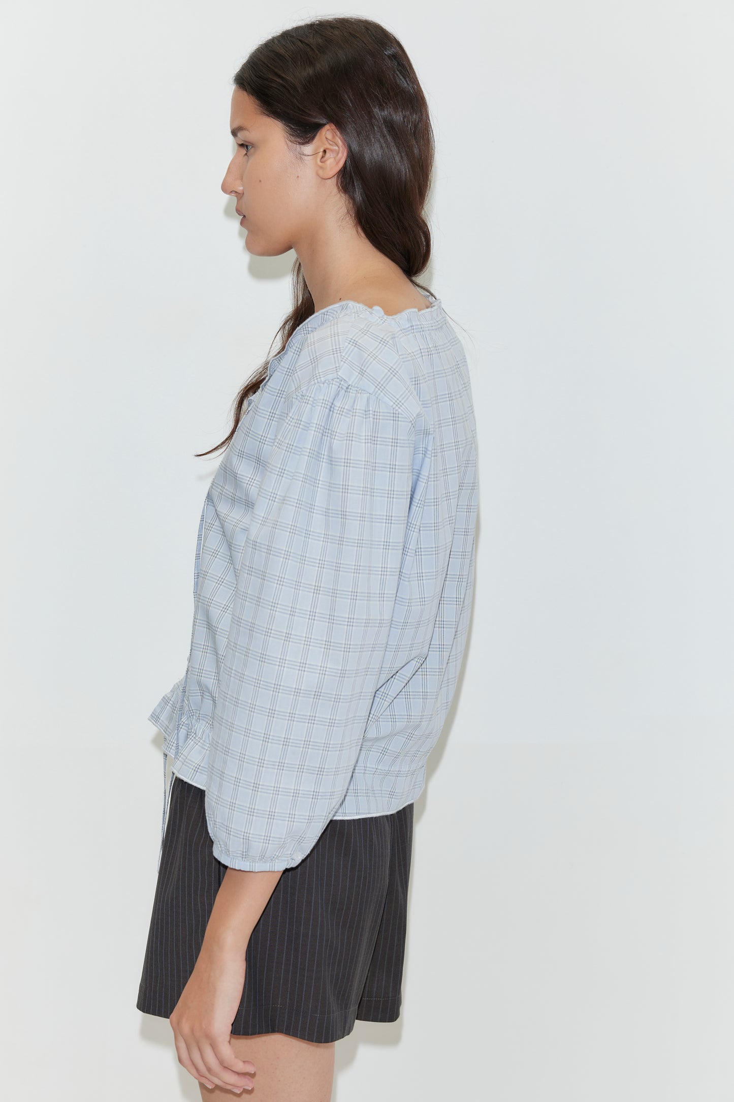 A woman with long brown hair stands sideways against a plain white background, wearing the Corded Top - Powder Check by Deiji Studios with puffed sleeves and dark pinstriped shorts.