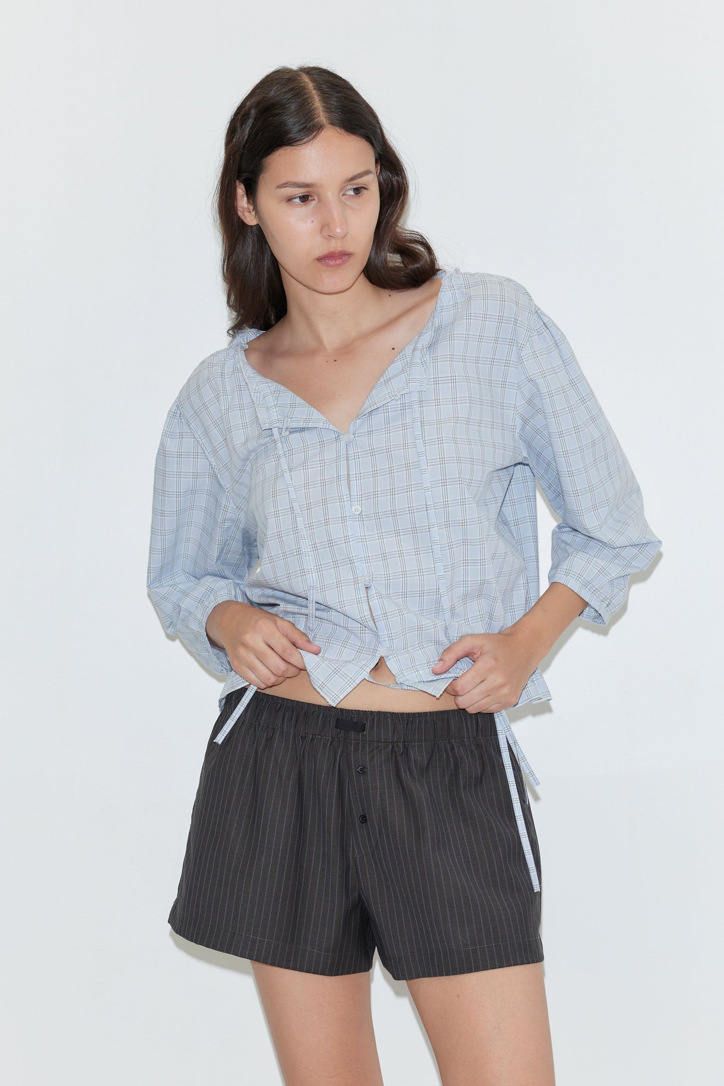 A woman with long brown hair wears the Corded Top - Powder Check by Deiji Studios and dark pinstriped shorts against a plain white background, holding the shirt’s hem with one hand and glancing slightly to the side.