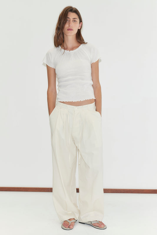 A person stands against a plain white background, wearing a fitted white short-sleeve top, Turn Pant - Off White with a loose wide-leg fit, and sandals, hands in their pockets and a neutral expression.