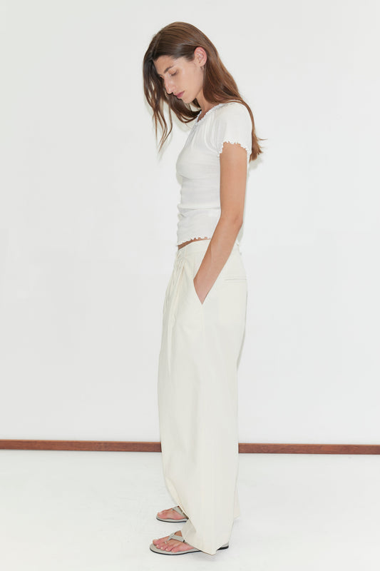 A woman with long brown hair stands sideways against a white background, wearing a white short-sleeve top, the Turn Pant - Off White from Deiji Studios, and sandals, with her hands in her pockets and head slightly bowed.