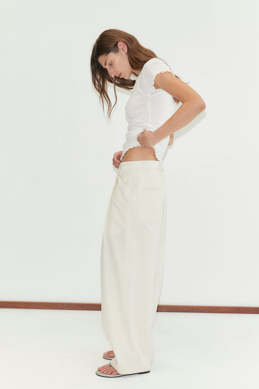 A woman stands sideways against a plain white background, adjusting her Turn Pant - Off White by Deiji Studios, paired with a white t-shirt.
