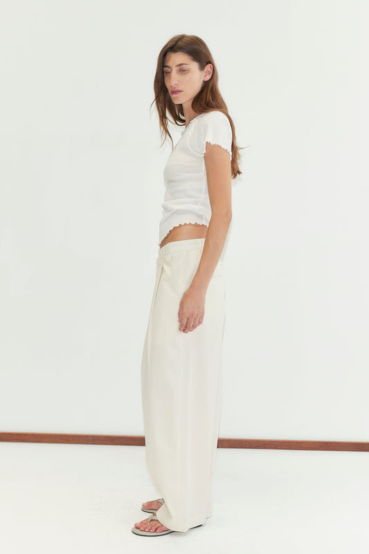 A woman with long brown hair stands sideways in a fitted white short-sleeve top, Deiji Studios’ Turn Pant in Off White, and sandals against a plain white background.