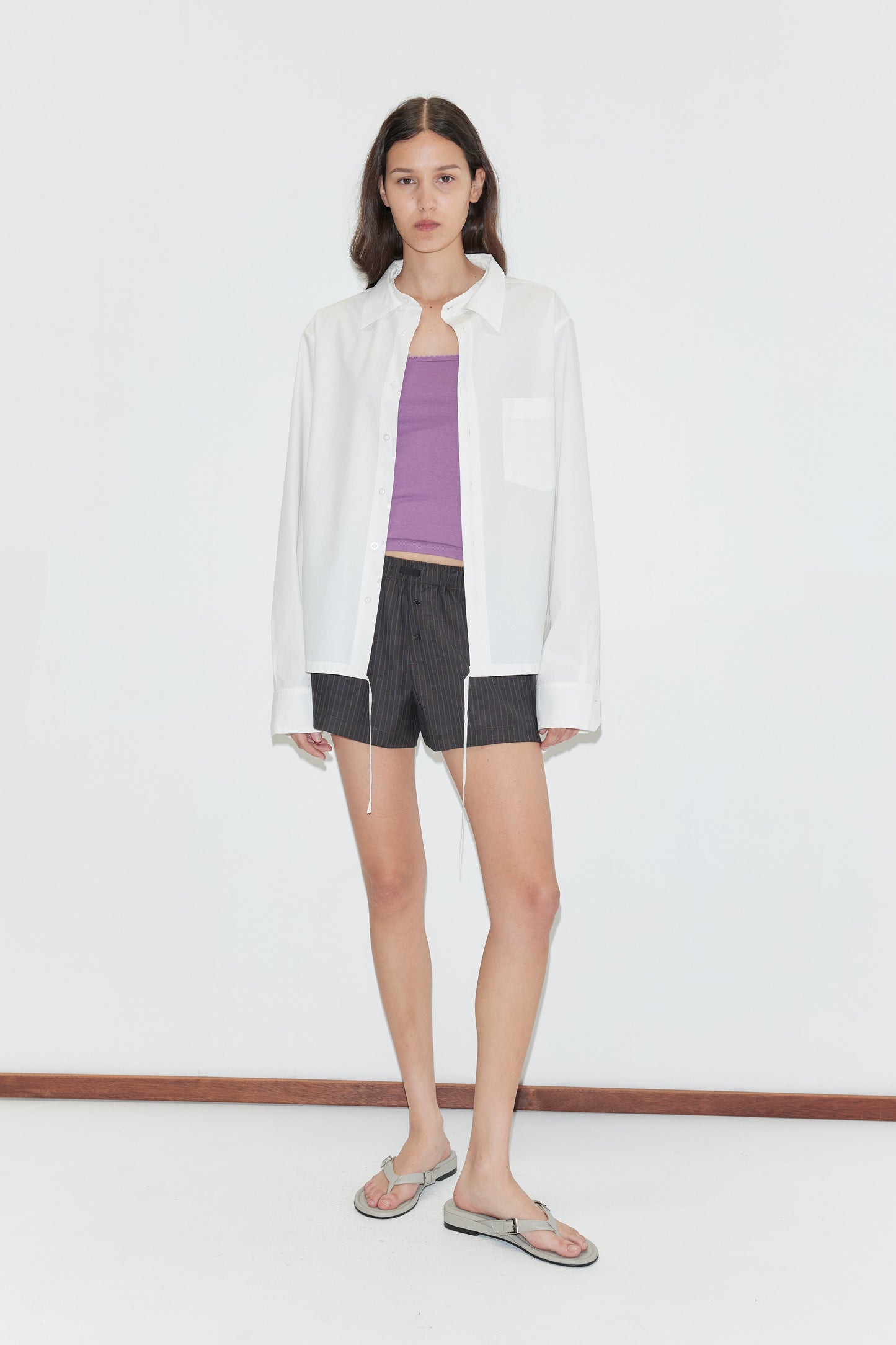 A woman with long brown hair and a neutral expression stands against a white background, wearing the Stand Shirt - White from Deiji Studios over a purple tank top, black pinstripe shorts, and silver flip-flops.