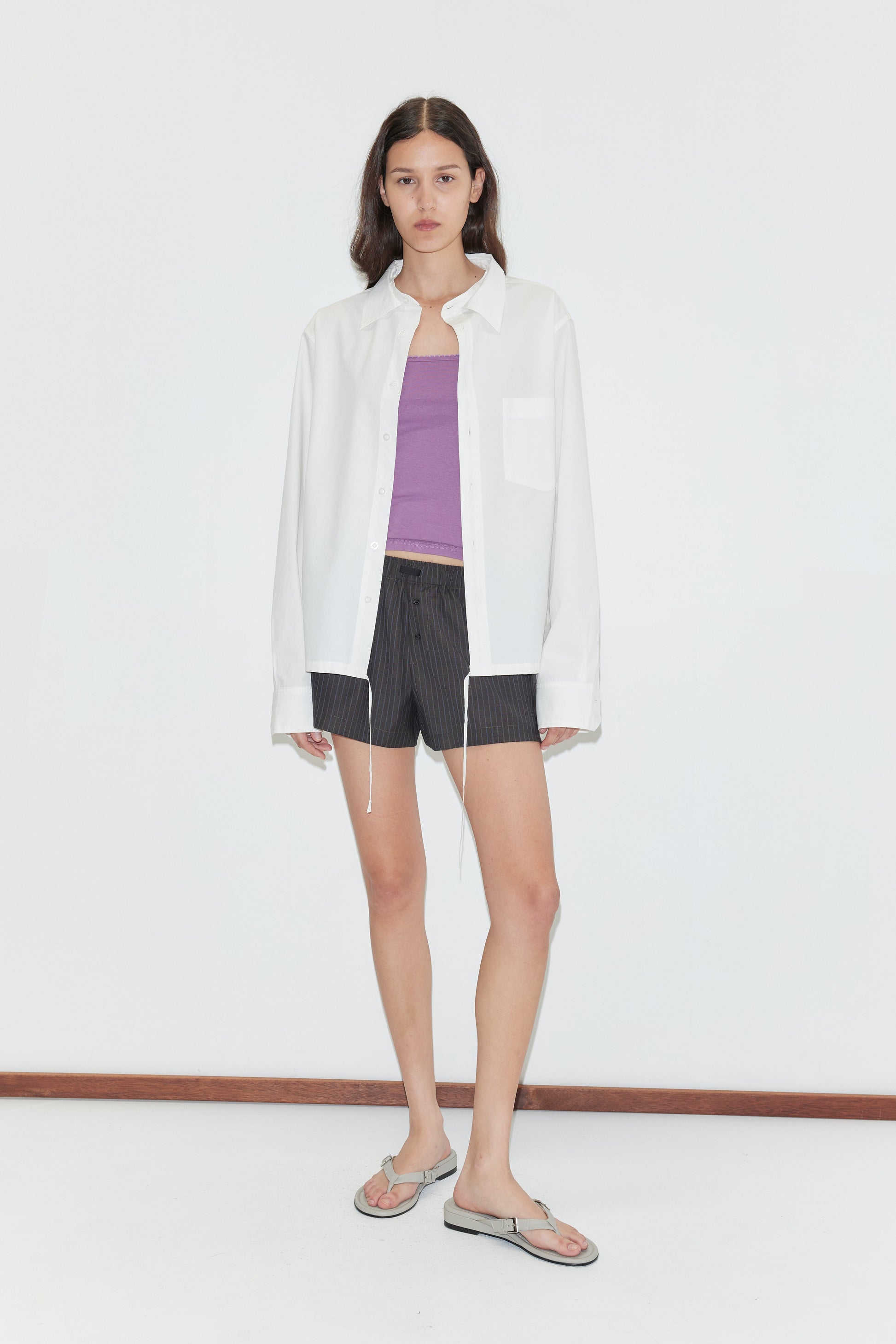 A woman with long brown hair and a neutral expression stands against a white background, wearing the Stand Shirt - White from Deiji Studios over a purple tank top, black pinstripe shorts, and silver flip-flops.