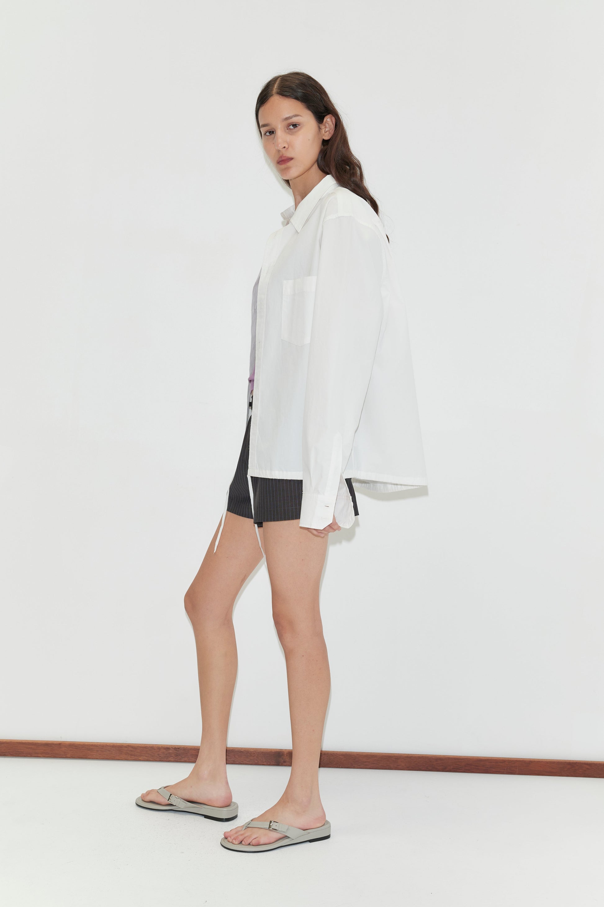 Against a plain white background, a person with long brown hair wears Deiji Studios' Stand Shirt - White with a mandarin collar, paired with dark shorts and gray sandals. They face slightly to the side with a neutral expression.