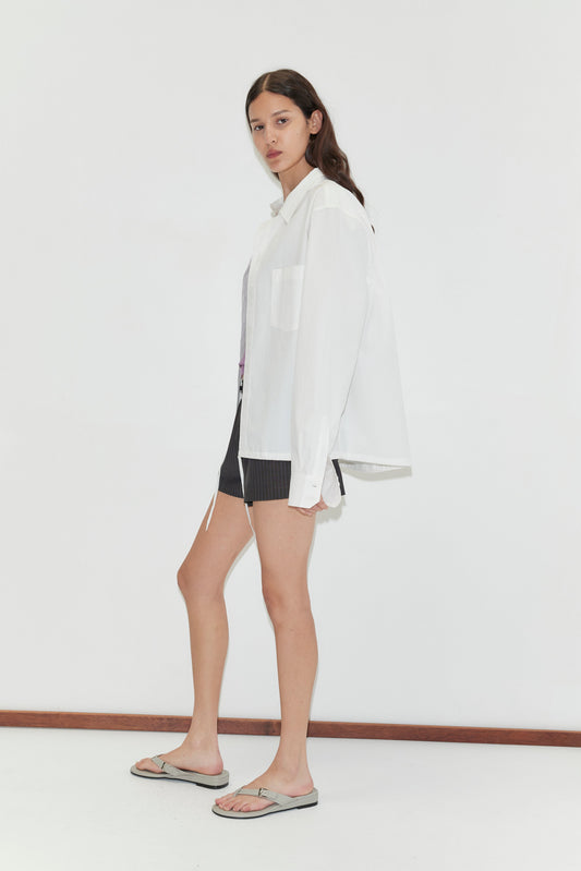 Against a plain white background, a person with long brown hair wears Deiji Studios' Stand Shirt - White with a mandarin collar, paired with dark shorts and gray sandals. They face slightly to the side with a neutral expression.