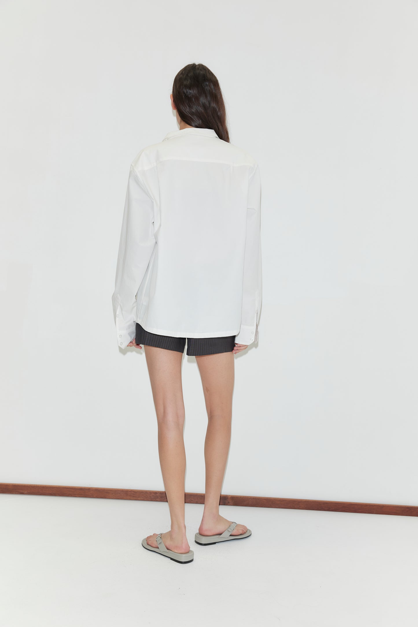A person with long dark hair stands facing away from the camera, wearing the Stand Shirt - White by Deiji Studios, paired with dark shorts and flip-flops, against a plain white background.