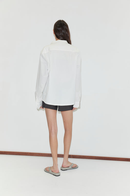 A person with long dark hair stands facing away from the camera, wearing the Stand Shirt - White by Deiji Studios, paired with dark shorts and flip-flops, against a plain white background.