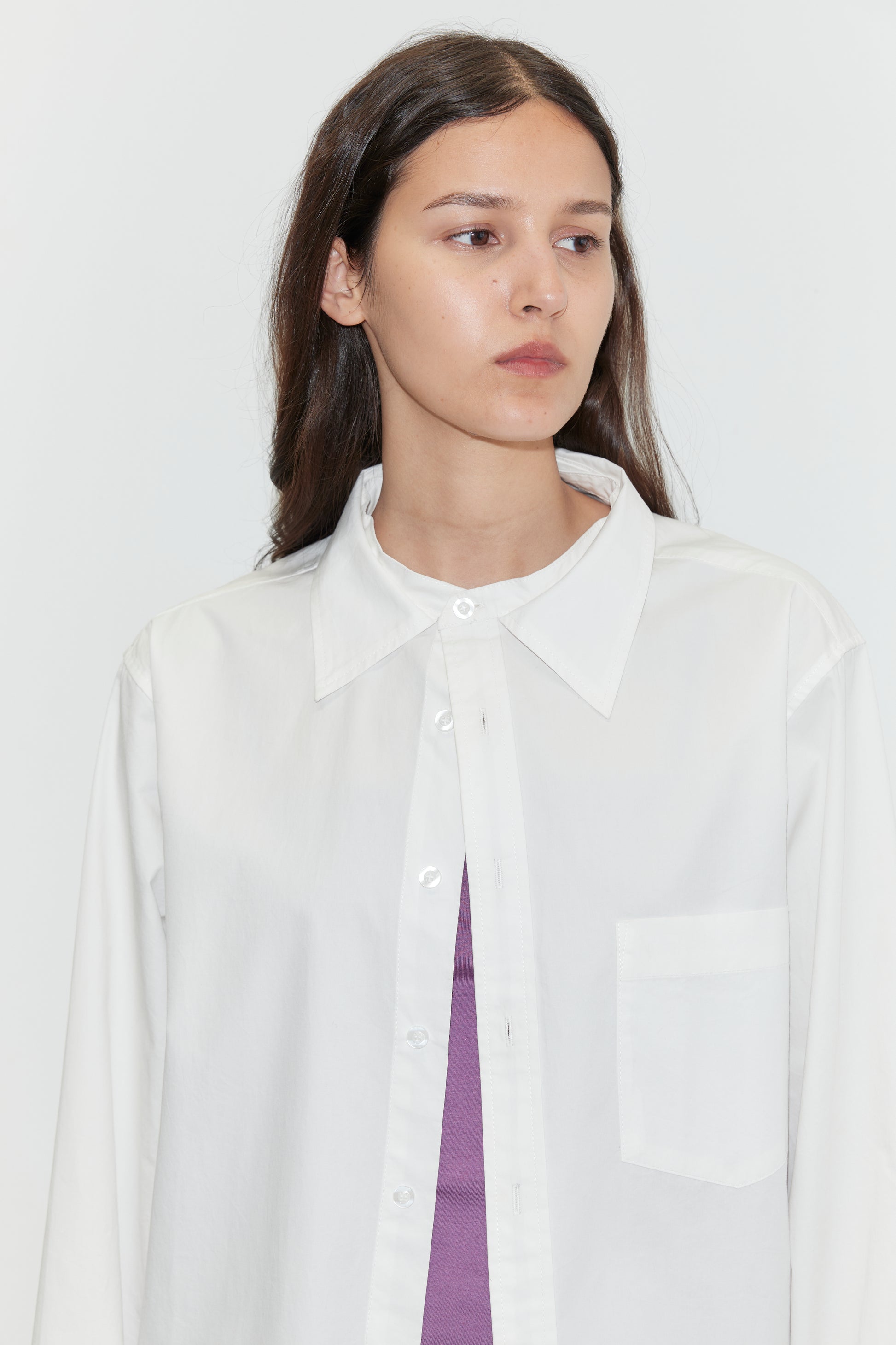 A woman with long brown hair wears the Stand Shirt - White from Deiji Studios over a purple top, standing against a plain white background and gazing slightly to the side.