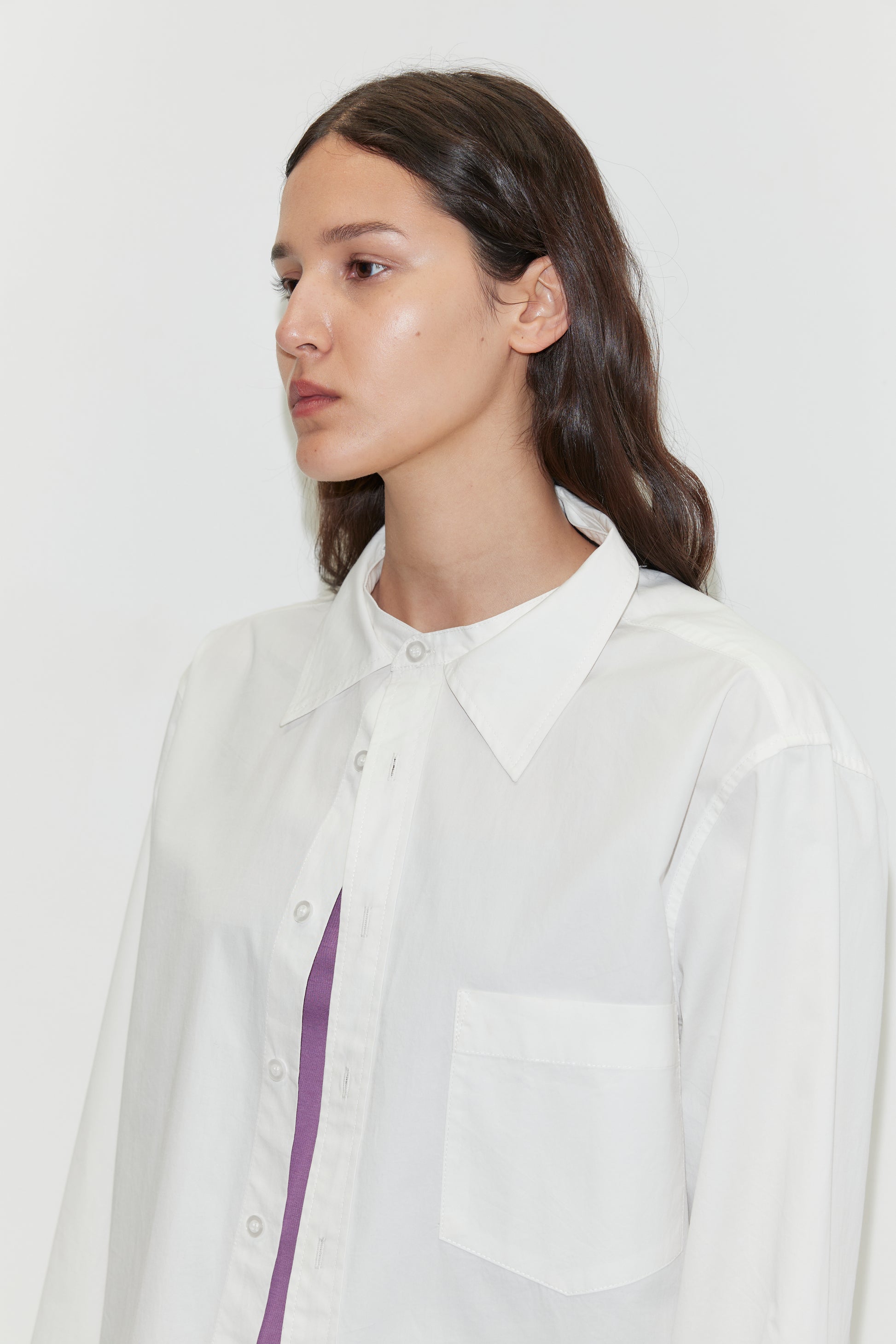 A person with long dark hair wears the Stand Shirt - White by Deiji Studios, an oversized cotton shirt with a mandarin collar, over a purple top while looking to the side against a plain white background.