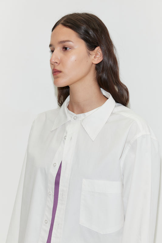 A person with long dark hair wears the Stand Shirt - White by Deiji Studios, an oversized cotton shirt with a mandarin collar, over a purple top while looking to the side against a plain white background.