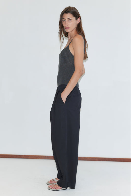 A woman with long brown hair, wearing a dark gray tank top, flip-flops, and the Turn Pant - Black by Deiji Studios in organic cotton, stands sideways against a plain white background, looking toward the camera.