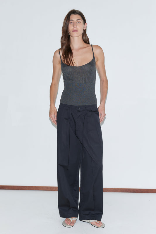 A person with long brown hair wears a fitted dark gray tank, Deiji Studios' Turn Pant in black organic cotton, and sandals, standing against a plain white background.