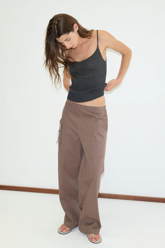 A woman with long brown hair wears the Adjust Pocket Pant - Sawdust and a black sleeveless top. Barefoot on a white floor, she looks down, adjusting her waistband against a plain white background.
