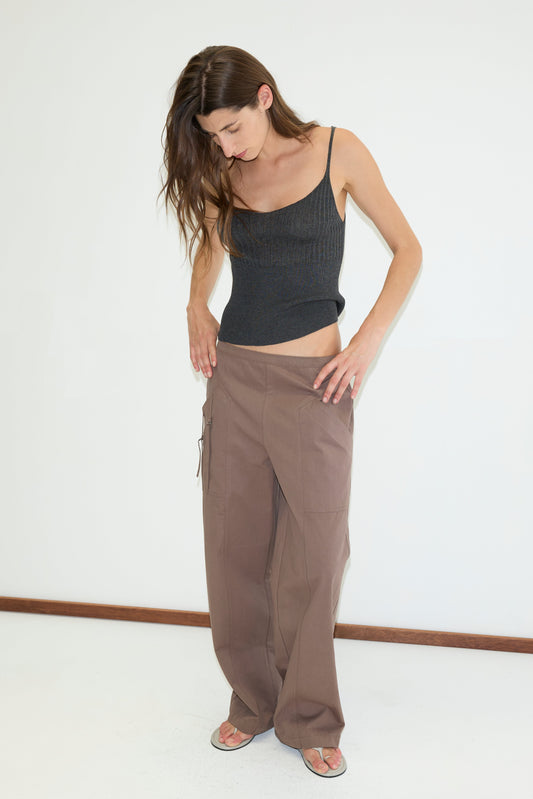 A woman with long brown hair in a black spaghetti strap top, relaxed mid-rise trousers, and sandals stands indoors against a white wall, looking down as she adjusts her Adjust Pocket Pant - Sawdust.