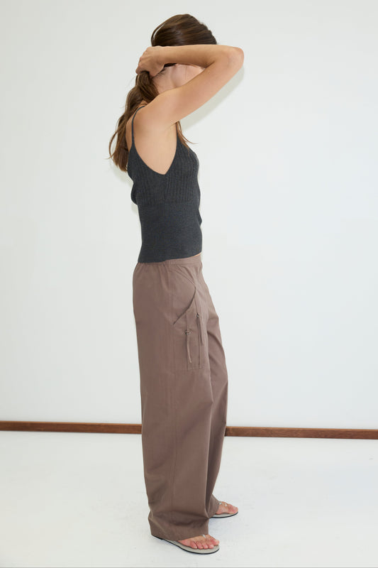 A woman stands sideways against a plain white wall, wearing the Adjust Pocket Pant in Sawdust with a black sleeveless top. She holds her hair back with both hands, partially covering her face.