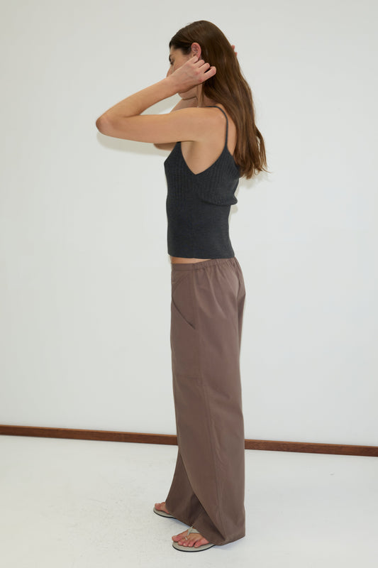 A woman with long brown hair stands in profile against a white wall, wearing a dark sleeveless top and the Adjust Pocket Pant - Sawdust, a relaxed mid-rise trouser made from organic cotton, with one hand at her ear and the other behind her head.