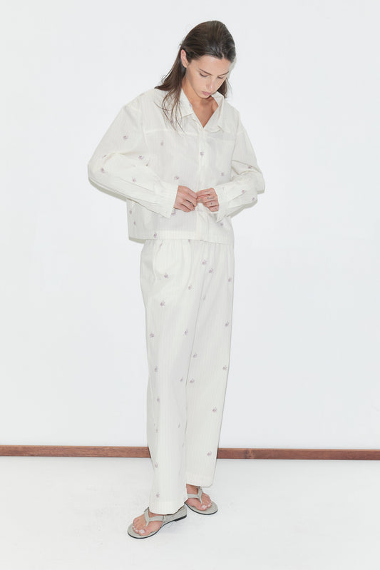 Wearing the Ease Trouser - Floral Stripe set, a person stands indoors adjusting the jacket. The white pajama ensemble features a loose fit and subtle floral stripe pattern, with straight leg pants enhancing its relaxed silhouette. White flip-flops complete the look against a minimal background.