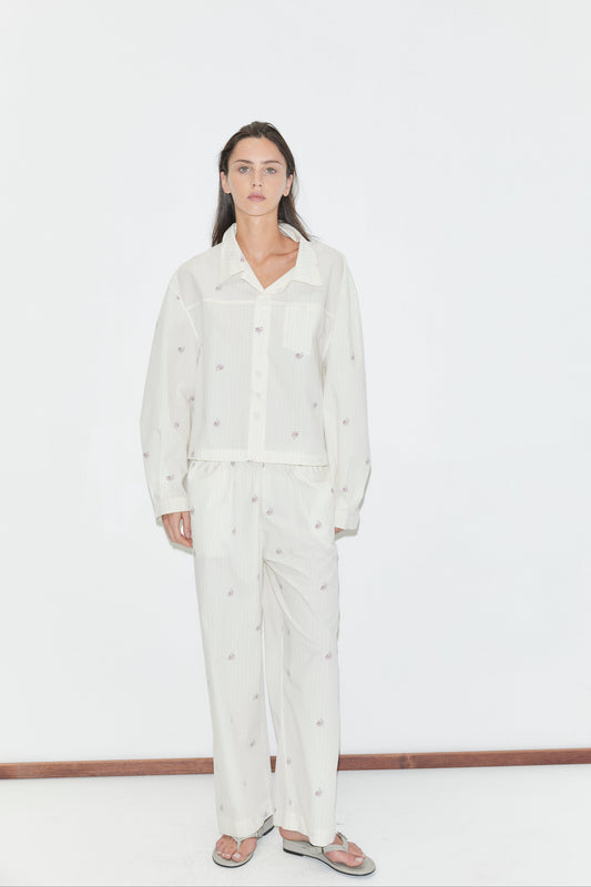 A person stands against a white background wearing loose, light-colored pajamas featuring the "Ease Trouser - Floral Stripe"—easy, straight leg trousers with a subtle floral stripe pattern—paired with flip-flops. They have long brown hair and a neutral expression.