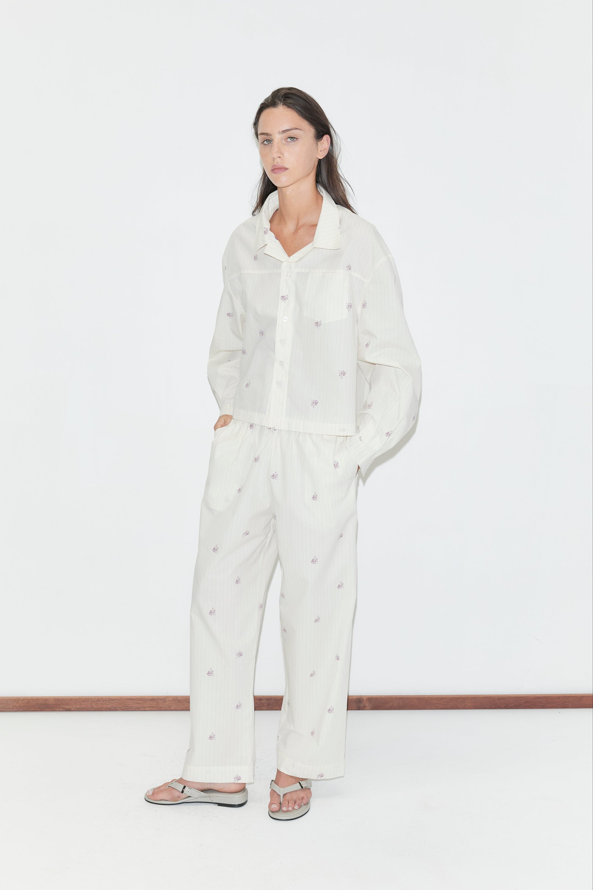 A woman stands against a white wall wearing loose, white button-up pajamas with a subtle dotted pattern and the Ease Trouser in Floral Stripe, hands in pockets, and gray flip-flops.