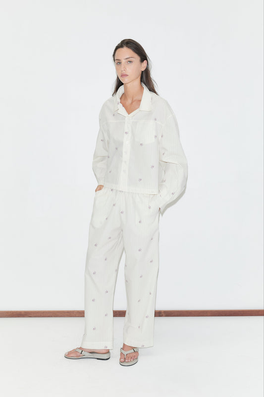 A woman stands against a white wall wearing loose, white button-up pajamas with a subtle dotted pattern and the Ease Trouser in Floral Stripe, hands in pockets, and gray flip-flops.