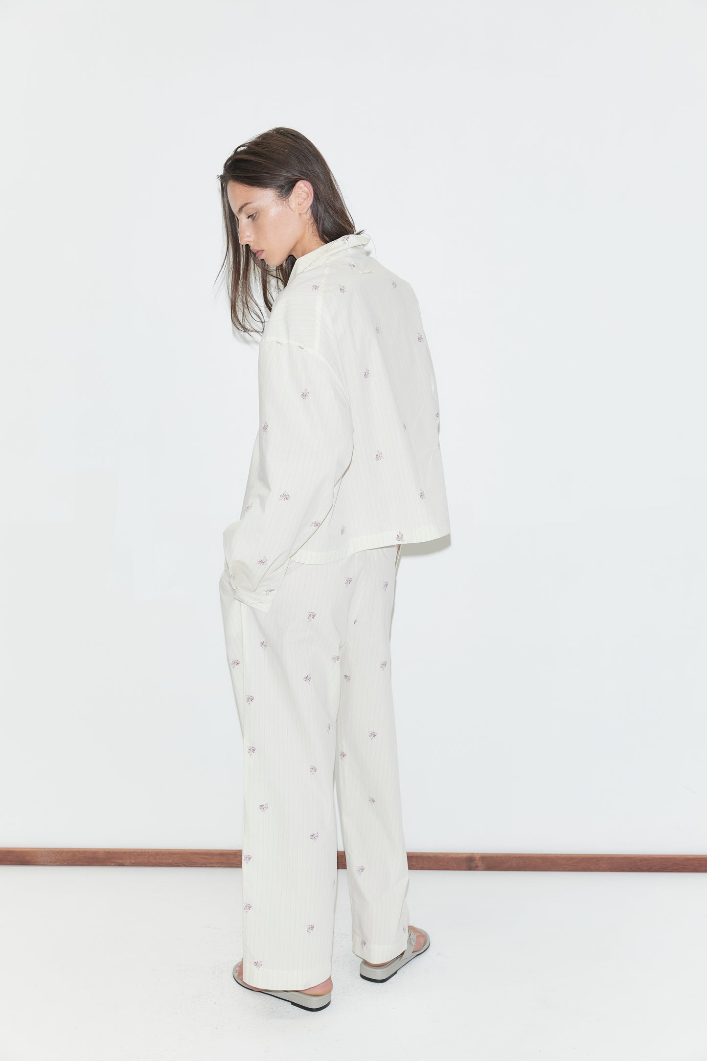 A person stands sideways against a white background, wearing the Ease Trouser - Floral Stripe pajama set, which features a loose fit with straight-leg trousers adorned with small, subtle floral stripe patterns. Their arms hang relaxed by their sides, and their long brown hair falls forward as they look down slightly.