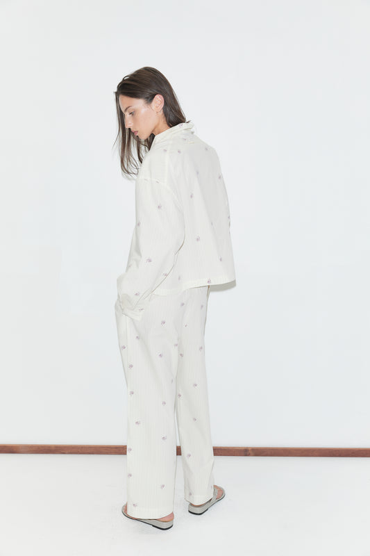 A person stands sideways against a white background, wearing the Ease Trouser - Floral Stripe pajama set, which features a loose fit with straight-leg trousers adorned with small, subtle floral stripe patterns. Their arms hang relaxed by their sides, and their long brown hair falls forward as they look down slightly.