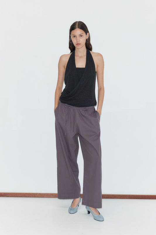 A woman with long dark hair stands against a white wall, wearing the Loop Pant in Mulberry Stripe, a sleeveless black halter top, and light blue heels. Her hands are in her pockets.
