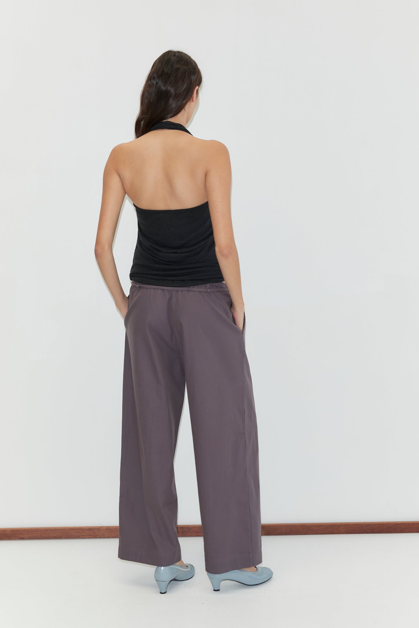 A woman faces away from the camera, wearing a black halter top, Loop Pant - Mulberry Stripe, and light blue heels against a plain white background.