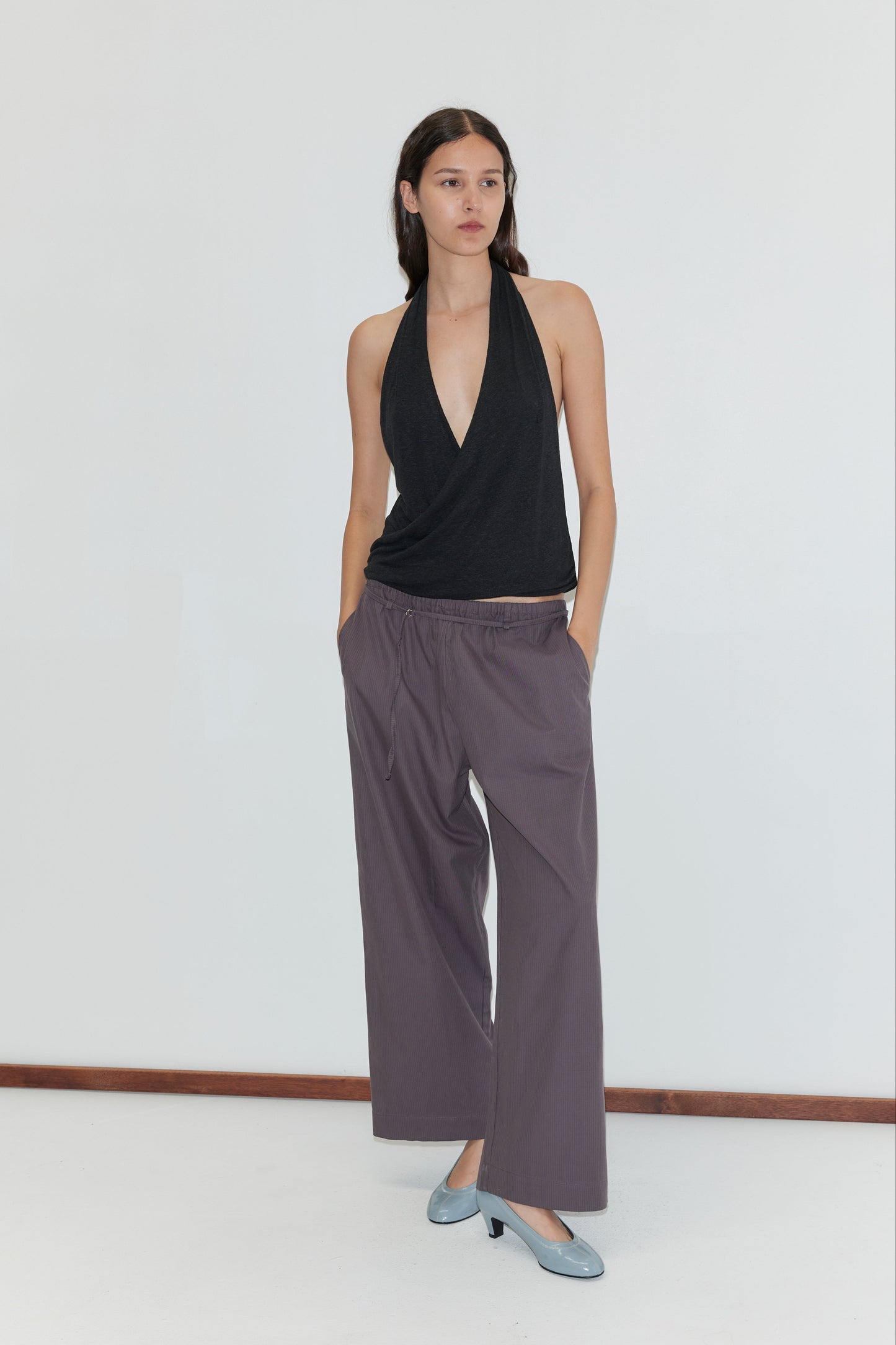 A woman with long dark hair and a neutral expression stands against a white wall, wearing a black halter top and Loop Pant - Mulberry Stripe with hands in pockets, paired with light blue low-heeled shoes.