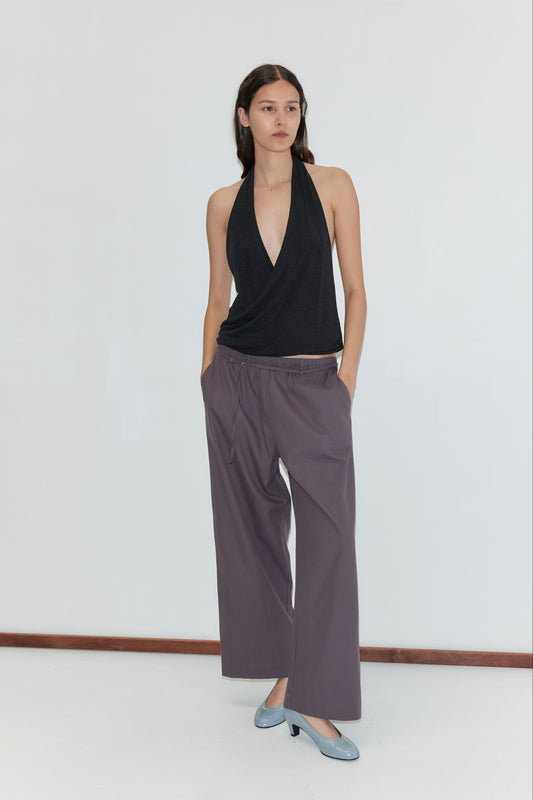 A woman with long dark hair and a neutral expression stands against a white wall, wearing a black halter top and Loop Pant - Mulberry Stripe with hands in pockets, paired with light blue low-heeled shoes.