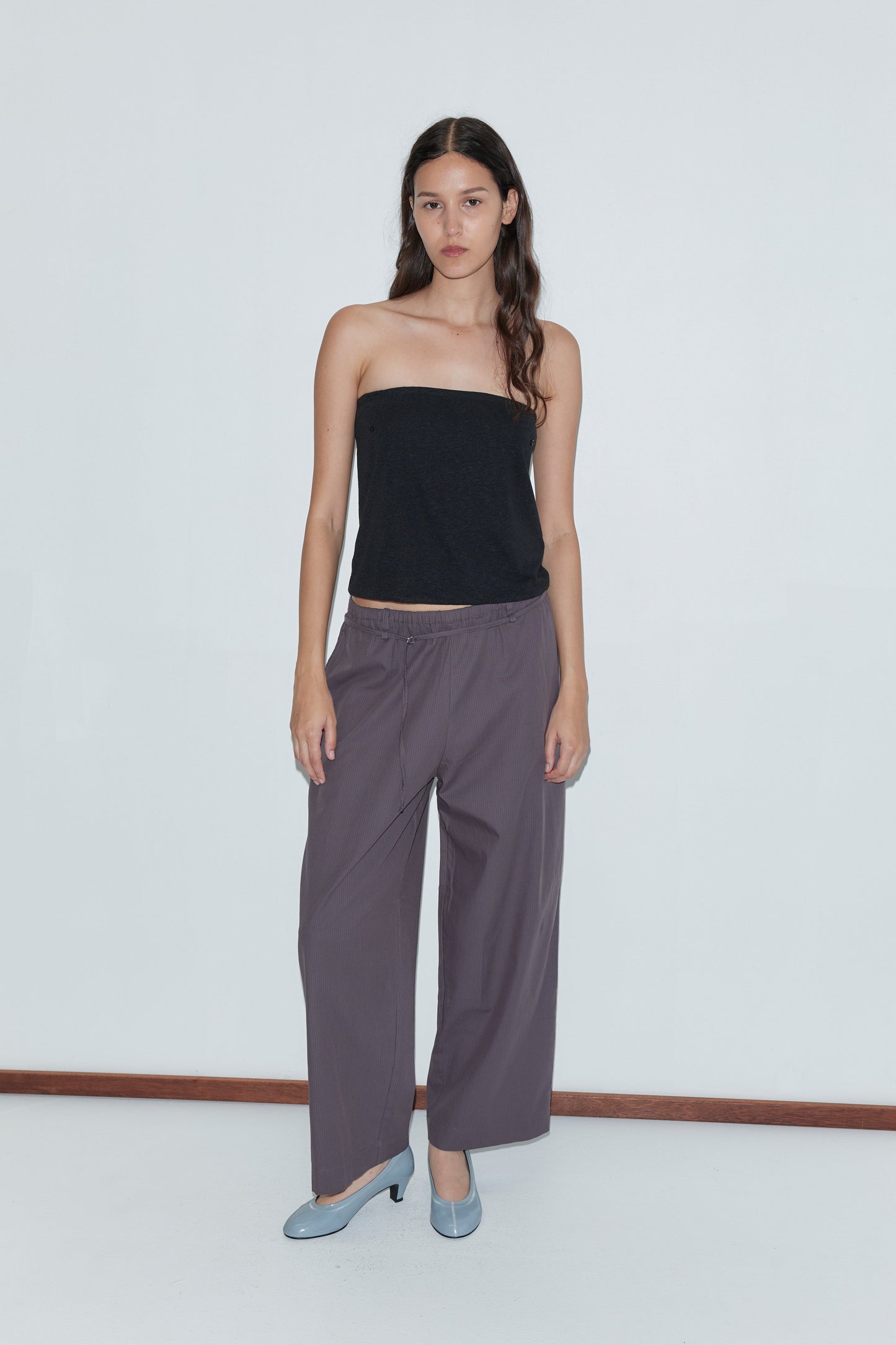 A woman with long dark hair wears a black strapless top, the Loop Pant - Mulberry Stripe, and light blue heels, standing against a plain white background.