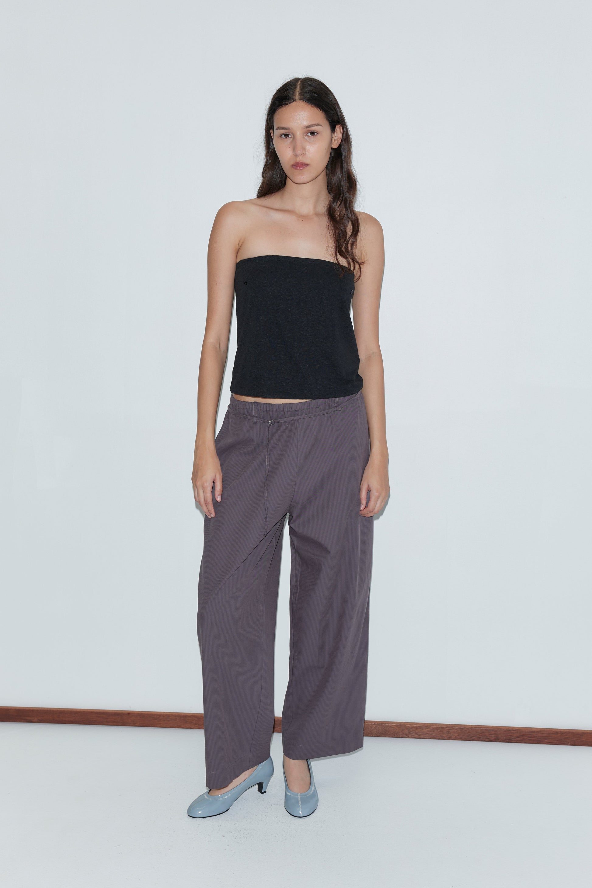 A woman with long dark hair wears a black strapless top, the Loop Pant - Mulberry Stripe, and light blue heels, standing against a plain white background.