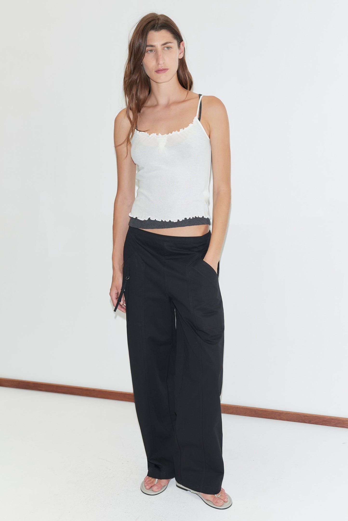 A woman with long brown hair and a neutral expression stands against a white background wearing a white camisole, the Deiji Studios Adjust Pocket Pant - Black, with one hand in her pocket and flip-flops.