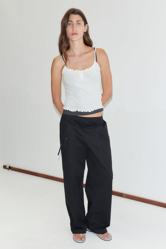 A person with long brown hair stands against a white background, wearing a Deiji Studios organic cotton camisole top, Adjust Pocket Pant - Black, and sandals. They face the camera with a neutral expression.