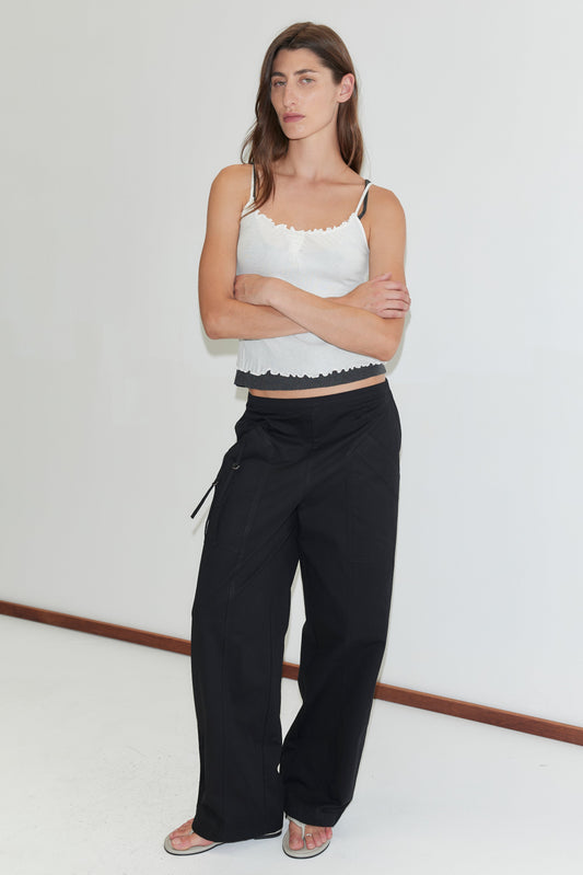 A woman with long brown hair stands against a white wall, wearing black Deiji Studios Adjust Pocket Pants, a lace-trim tank top, and sandals. She has her arms crossed and maintains a neutral expression.
