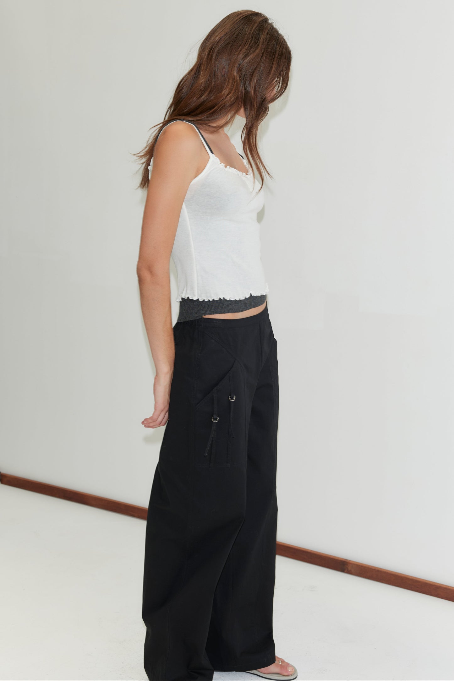 A woman with long brown hair stands indoors, facing away and looking down. She wears a white tank top and Deiji Studios' Adjust Pocket Pant in black with large pockets, set against a plain white background.