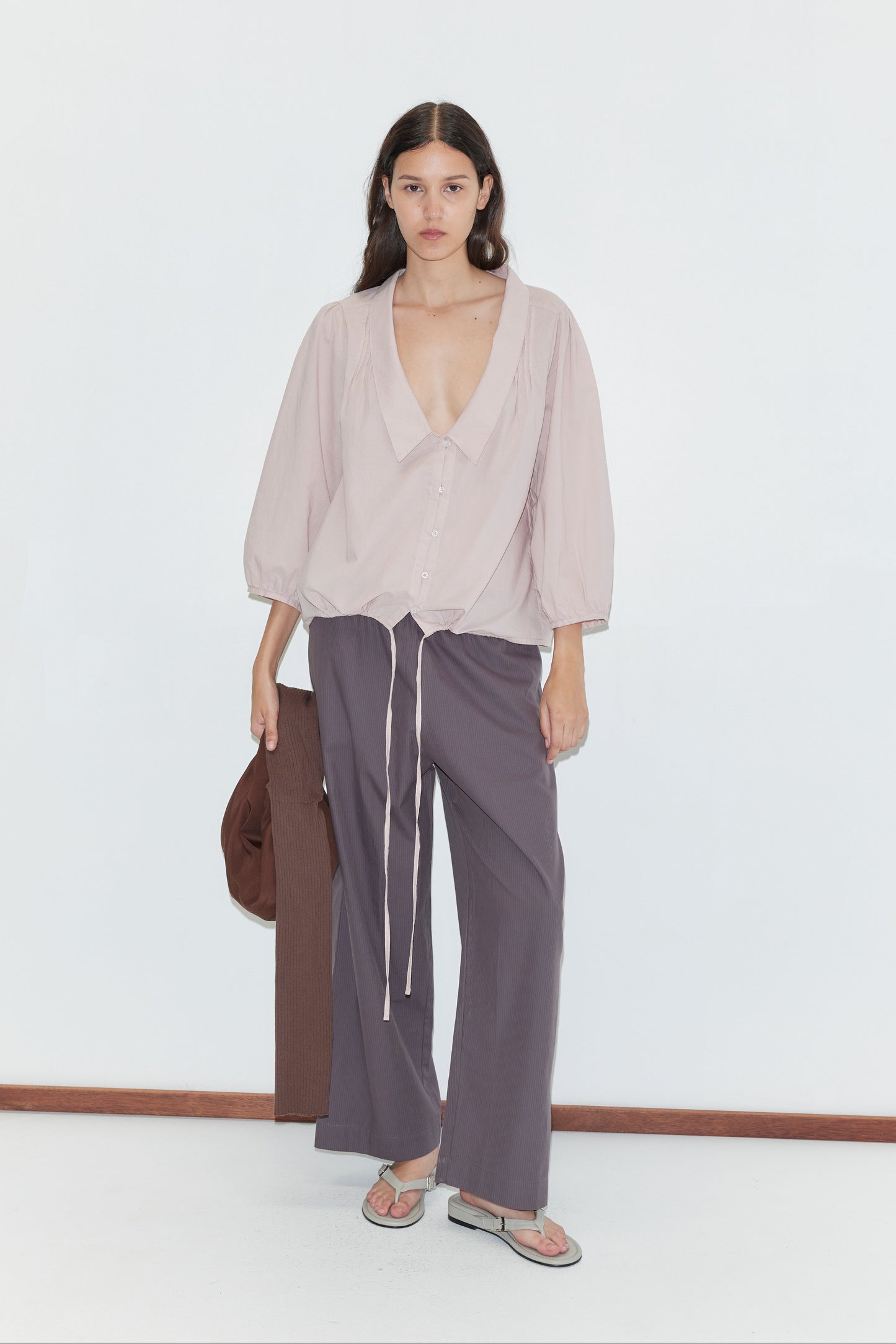 A woman with long, wavy dark hair and a neutral expression stands against a white background, wearing a light pink blouse, purple-gray pants, flip-flops, holding a brown jacket, and carrying the Hazel Knit Strap Bag.
