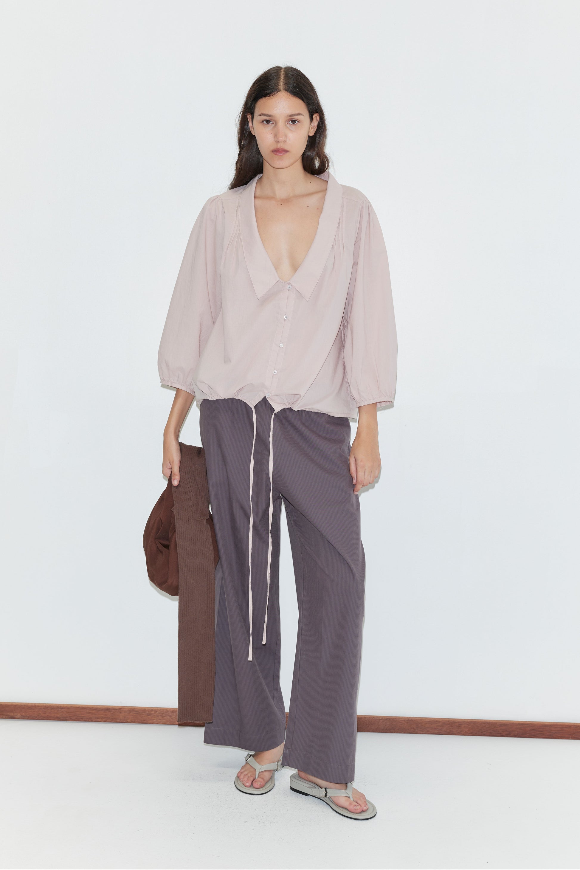 A woman with long, wavy dark hair and a neutral expression stands against a white background, wearing a light pink blouse, purple-gray pants, flip-flops, holding a brown jacket, and carrying the Hazel Knit Strap Bag.