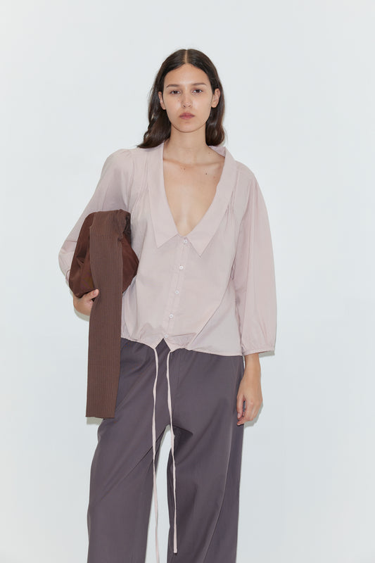 A woman with long dark hair wears a loose, light mauve blouse and dark pants, holding a brown garment over her arm. She accessorizes with the Knit Strap Bag - Hazel and stands against a plain white background.