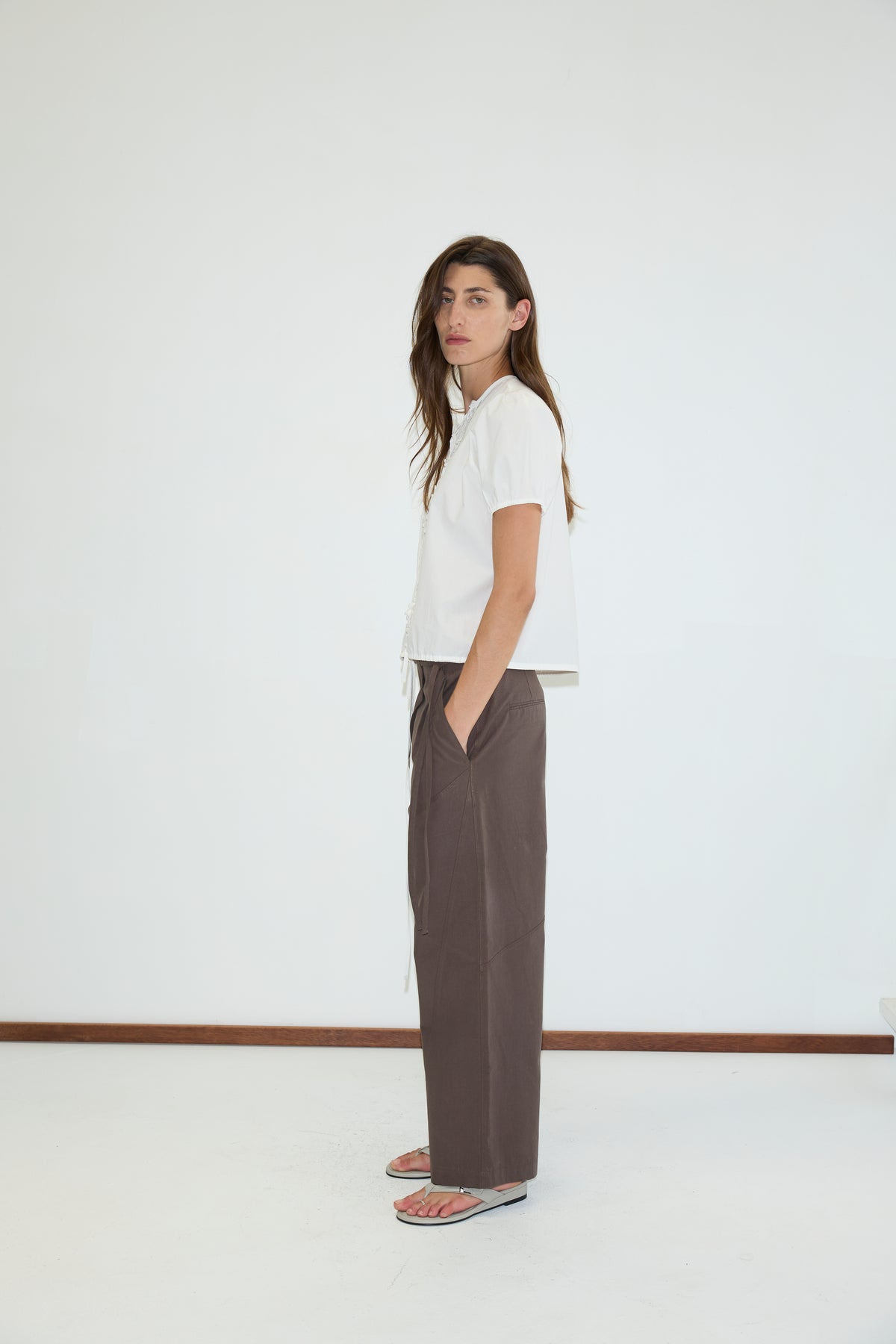 A person with long hair stands sideways, wearing a white short-sleeve shirt, Flag Tie Pant in Dark Brown by Deiji Studios, and sandals. The background is plain white with a brown floor trim.