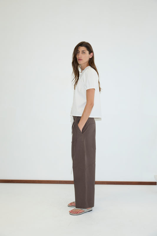 A person with long hair stands sideways, wearing a white short-sleeve shirt, Flag Tie Pant in Dark Brown by Deiji Studios, and sandals. The background is plain white with a brown floor trim.