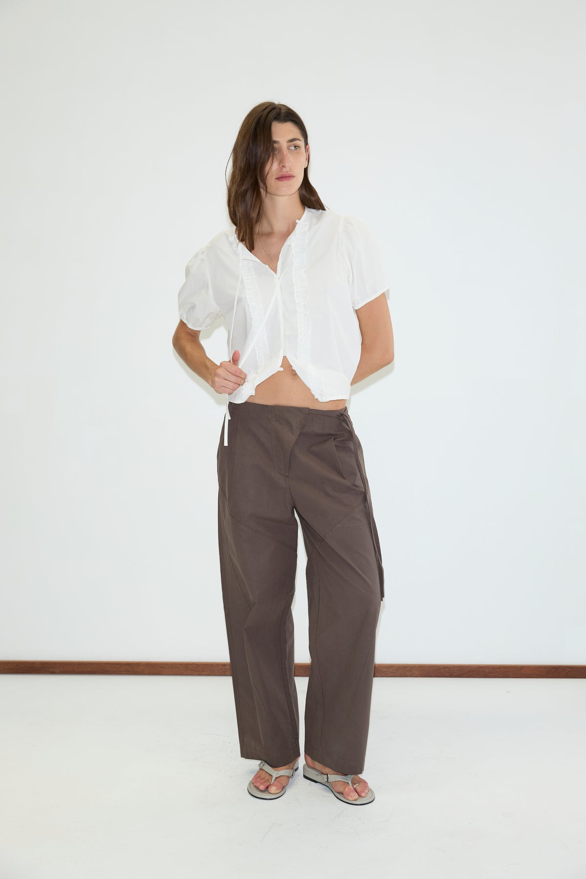 A person stands indoors wearing a white short-sleeve blouse, Deiji Studios Flag Tie Pant in dark brown, and sandals against a plain white wall.
