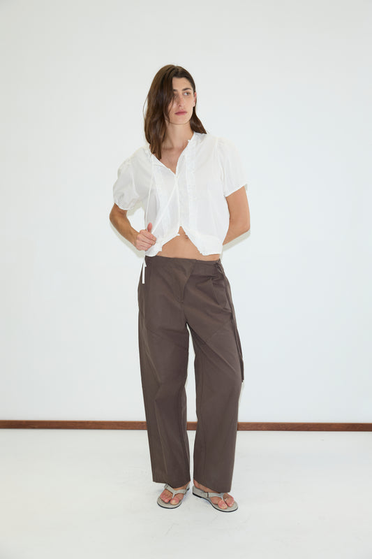 A person stands indoors wearing a white short-sleeve blouse, Deiji Studios Flag Tie Pant in dark brown, and sandals against a plain white wall.