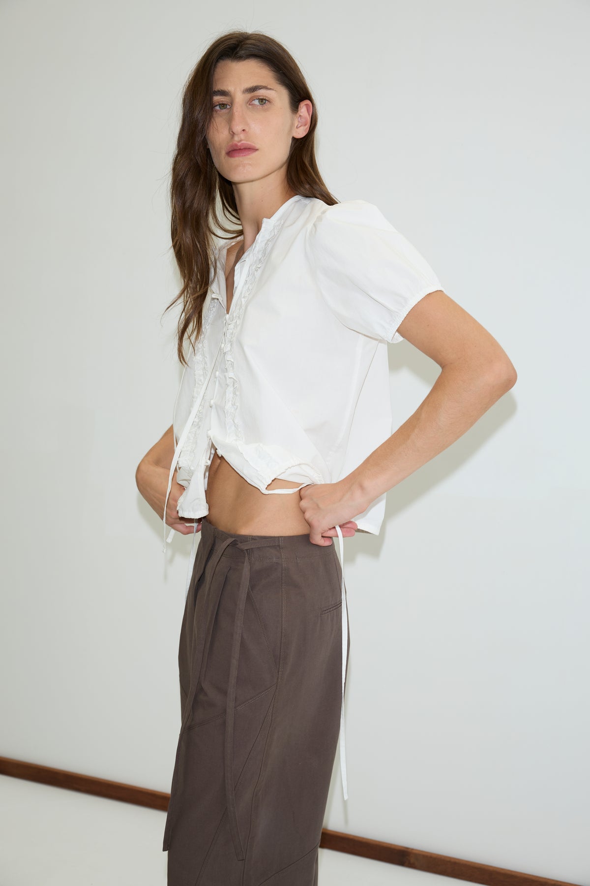A person with long brown hair stands against a plain white wall, wearing a white short-sleeve blouse and high-waisted Deiji Studios Flag Tie Pant in dark brown, looking confidently at the camera.