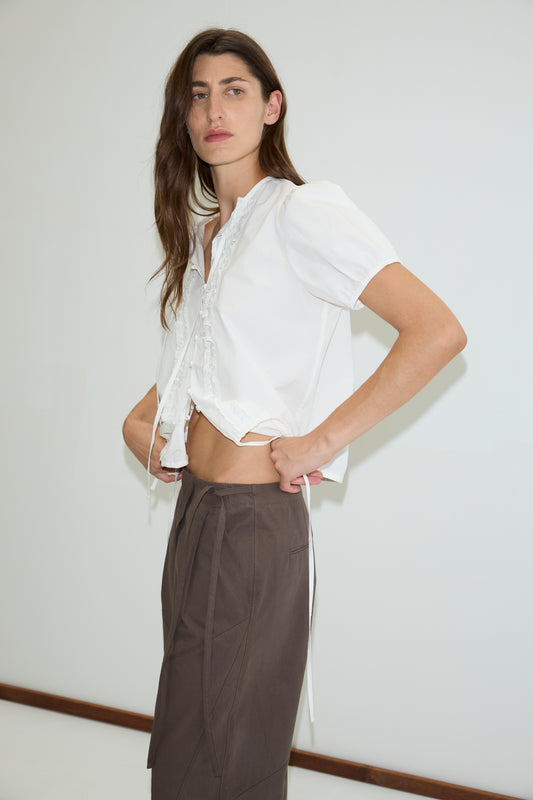 A person with long brown hair stands against a plain white wall, wearing a white short-sleeve blouse and high-waisted Deiji Studios Flag Tie Pant in dark brown, looking confidently at the camera.