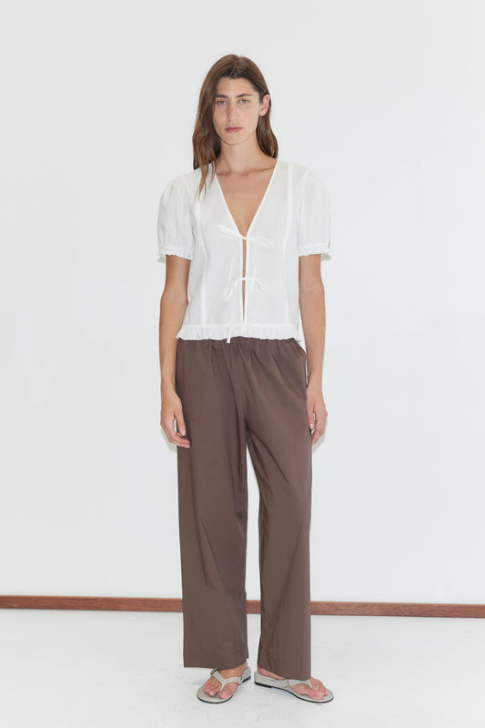 A person with long brown hair and a neutral expression stands against a white wall wearing a sheer white short-sleeve blouse, the Ease Trouser - Chocolate by Deiji Studios in organic cotton, and flat sandals.