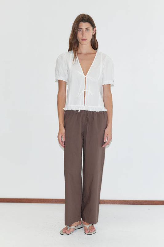 A person stands against a white background, wearing a Deiji Studios white short-sleeved tie-front blouse, Ease Trouser - Chocolate in organic cotton, and sandals. They have long hair and a neutral expression.